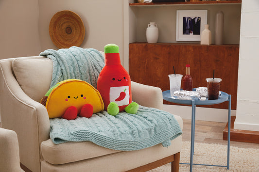 Taco and Hot Sauce Magnetic Plush (Extra Large) – Better Together sitting on a couch with a knitted blanket and a side table.