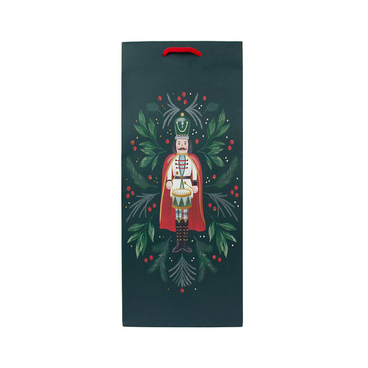 A dark teal Christmas-themed gift bag with a red handle, featuring an illustration of a nutcracker surrounded by green holly leaves and red berries, centered on a plain white background.