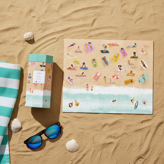Just Beachy 1000-piece jigsaw puzzle displayed on sandy beach, alongside packaging, sunglasses, and beach towel.