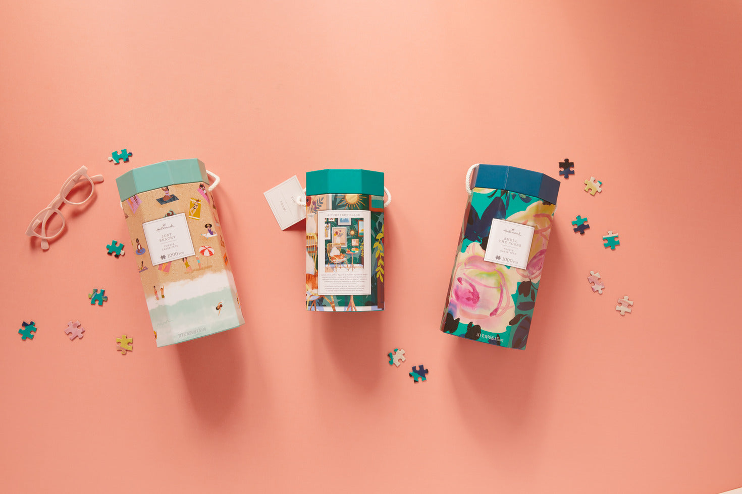 Jigsaw puzzles in decorative packaging with floral designs, displayed on a pastel pink background with puzzle pieces scattered.