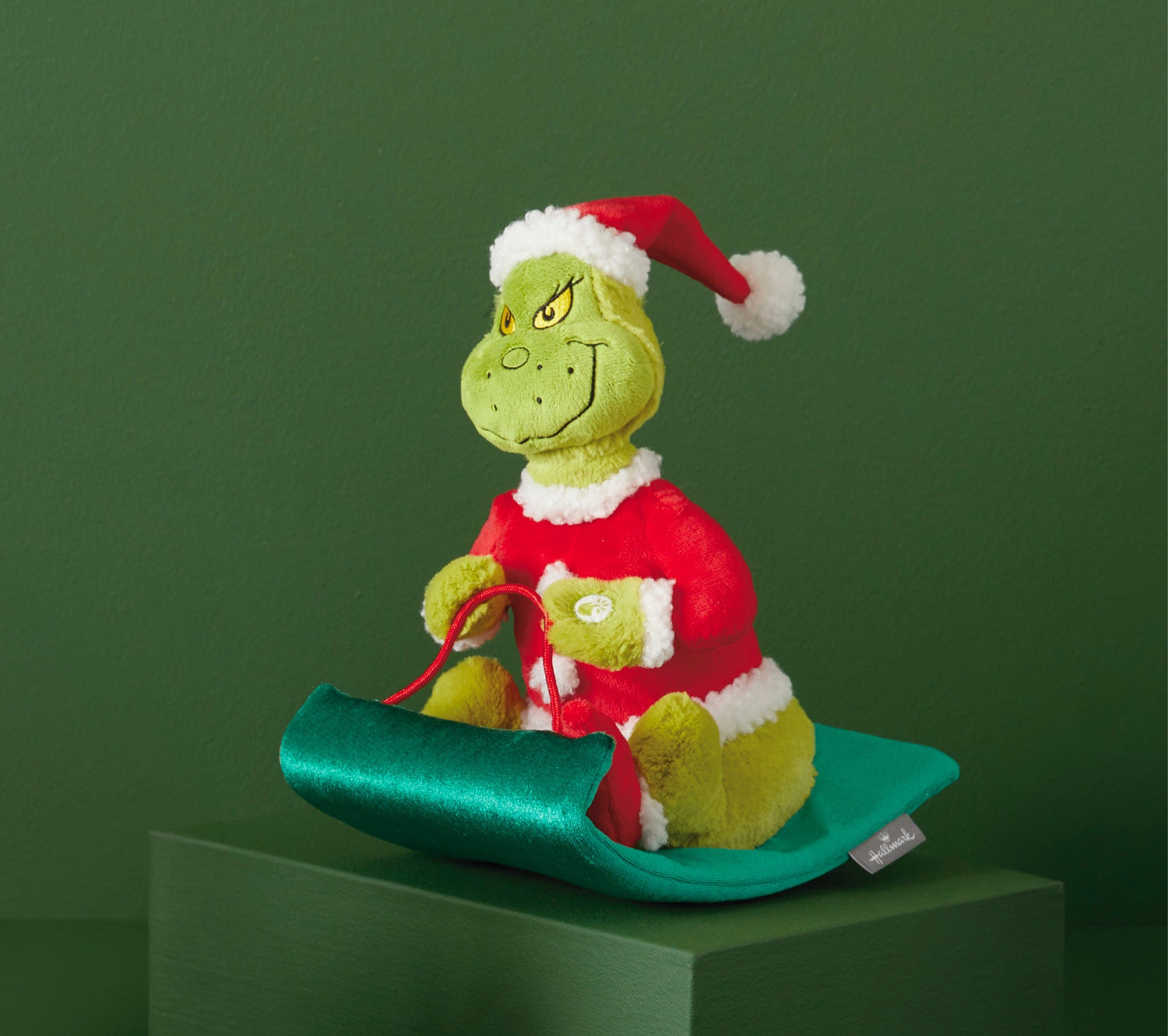 The Grinch plush toy in a Santa outfit, sitting on a green sled, detailed with bright colours and festive features.