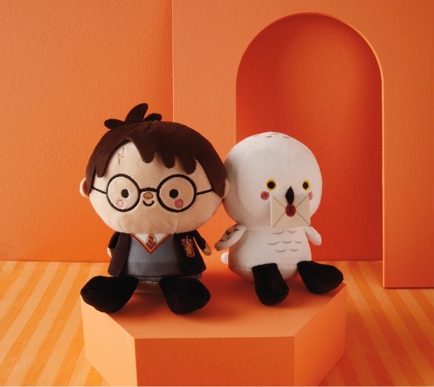 Harry Potter plush set featuring Harry and Hedwig, seated on a geometric pedestal with an orange backdrop.