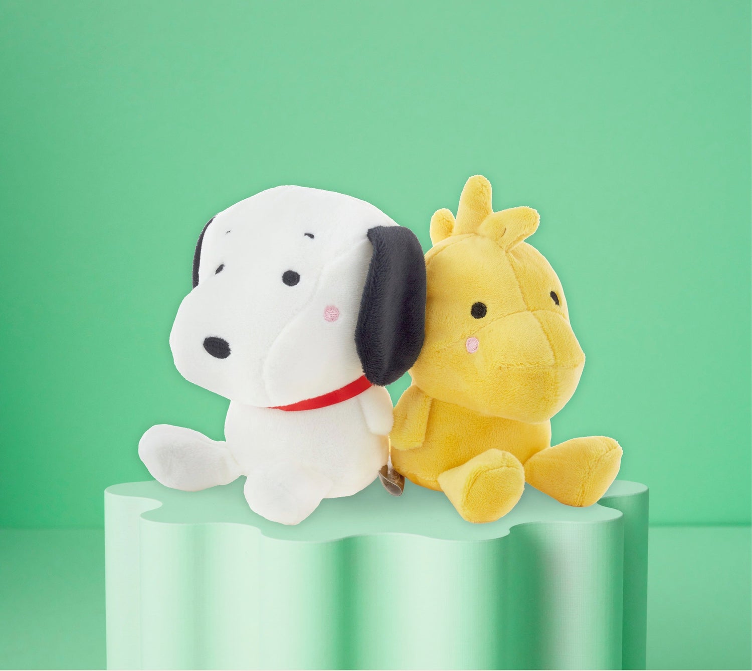 PEANUTS plush toys featuring Snoopy and Woodstock against a green background on a sculpted pedestal.