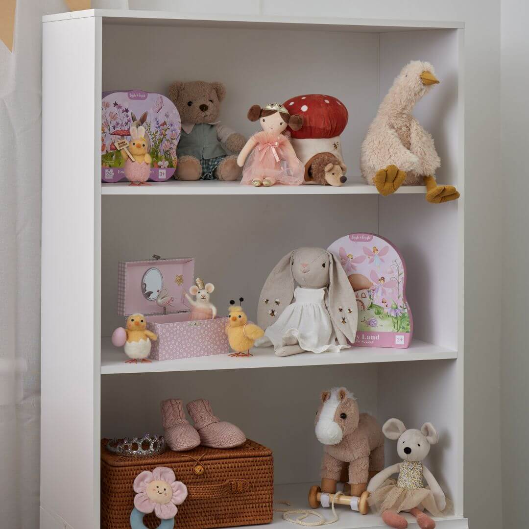 Wiggles the Duck soft toy among various plush toys on a white shelf, showcasing its cream colour and child-safe design.