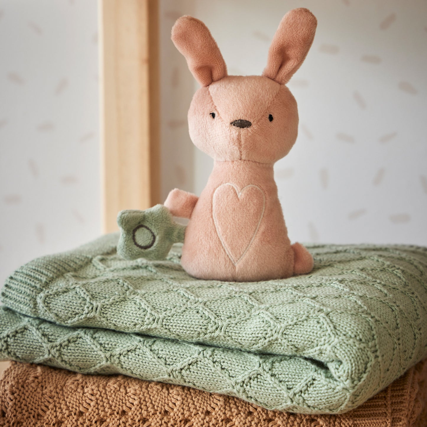 Freya Plush Bunny Rattle made from soft fabric, featuring a heart detail, displayed on a knitted green blanket.