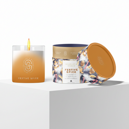A lit glass jar candle and its patterned cylindrical packaging container on a white pedestal. The candle is clear with a warm amber-to-white gradient wax and a visible flame.