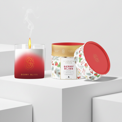 A lit scented candle in a clear glass jar with a red-to-white gradient and a gold logo, shown beside a floral-patterned tin and a red lid on a clean white studio set. Visible text includes 'GOLDEN GEM' and 'BERRY BLISS'.