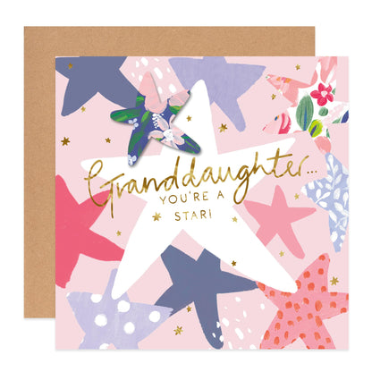A square greeting card with a kraft envelope behind it. The card has a pastel pink background with illustrated starfish in navy, pink, purple, and coral, plus small gold stars and the text 'Granddaughter.