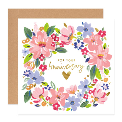A square anniversary greeting card with a white front, featuring a colorful floral wreath illustration in pink, blue, green, and red, with gold script text reading 'FOR YOUR Anniversary' and a gold heart. A kraft brown envelope is placed behind the card on a white background.