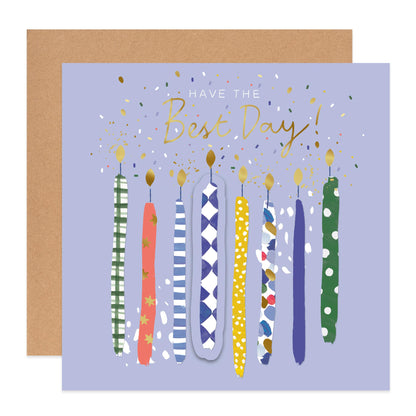 A square birthday card with a lavender background, gold script text reading 'HAVE THE Best Day!', and nine colorful illustrated candles in different patterns. A plain brown envelope is partially visible behind the card on a white background.
