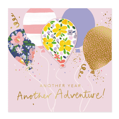 Square greeting card design with a light pink background, three illustrated balloons in floral and gold patterns, confetti, streamers, and the text 'ANOTHER YEAR. Another Adventure!'.