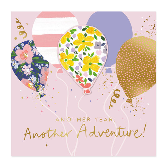Square greeting card design with a light pink background, three illustrated balloons in floral and gold patterns, confetti, streamers, and the text 'ANOTHER YEAR. Another Adventure!'.