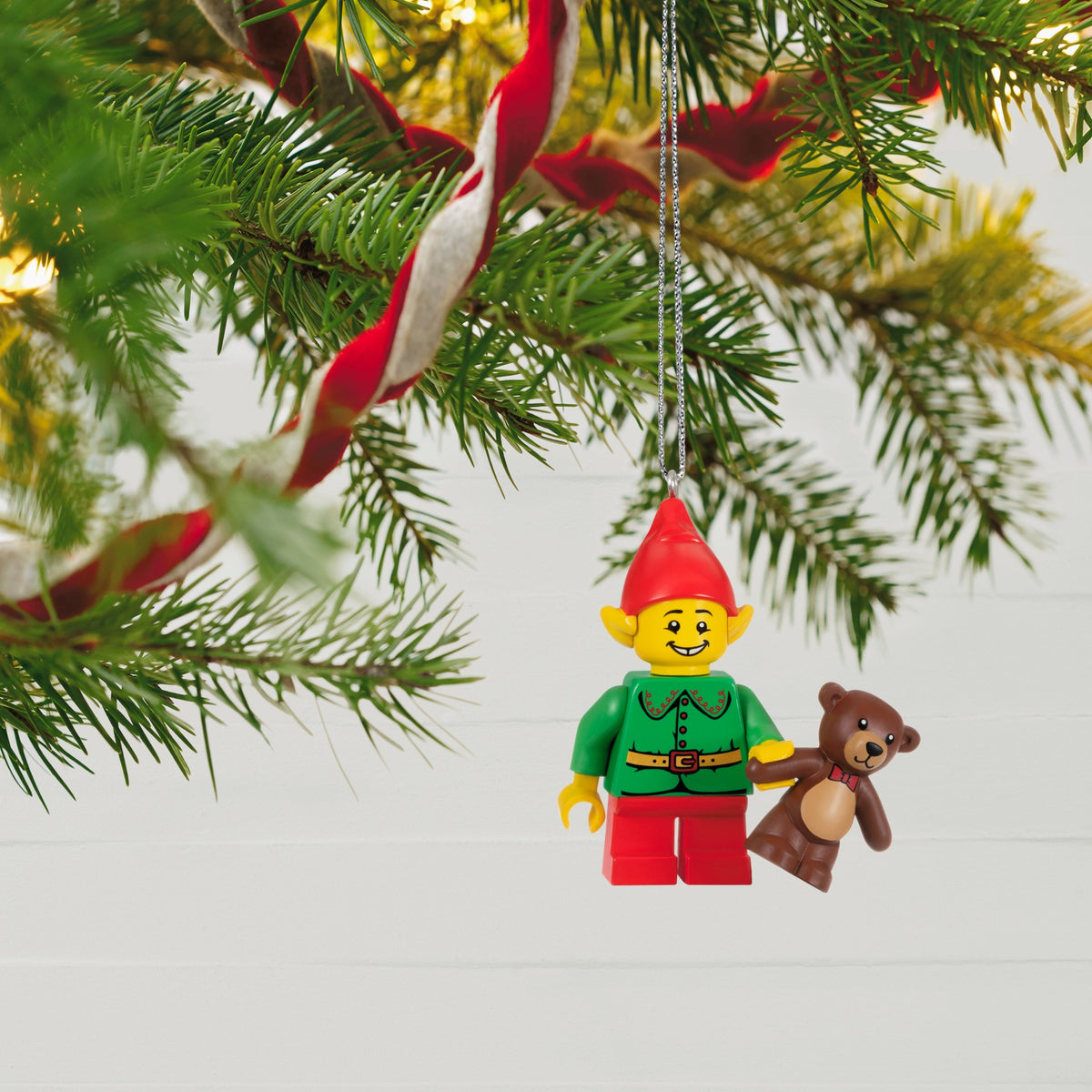 Christmas ornament of a LEGO-like elf holding a teddy bear hanging on a tree branch.