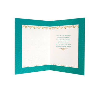 An open folded greeting card with a teal-green border and cream inner panels, featuring decorative gold foil accents and printed message text on the right side.