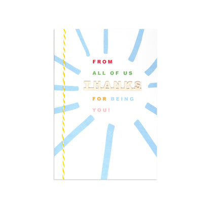 A square greeting card with a white background, featuring a light blue sunburst pattern and a yellow twisted thread spool graphic along the left edge. The text on the card reads 'FROM ALL OF US THANKS FOR BEING YOU!' in red, green, beige, and pink lettering.