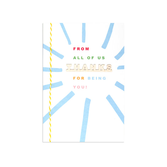 A square greeting card with a white background, featuring a light blue sunburst pattern and a yellow twisted thread spool graphic along the left edge. The text on the card reads 'FROM ALL OF US THANKS FOR BEING YOU!' in red, green, beige, and pink lettering.