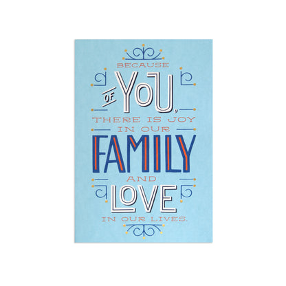 A square art print with a light blue background featuring a decorative typographic design that reads, 'BECAUSE OF YOU THERE IS JOY IN OUR FAMILY AND LOVE IN OUR LIVES.' The text uses white, navy blue, and orange-red lettering with small ornamental flourishes.