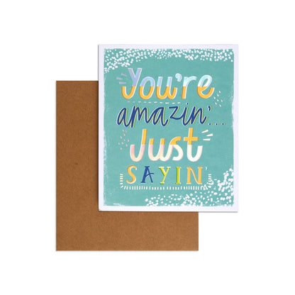 A square greeting card with a teal background and colorful hand-lettered text reading 'You're amazin' Just SAYIN', shown with a brown kraft envelope behind it on a white background.