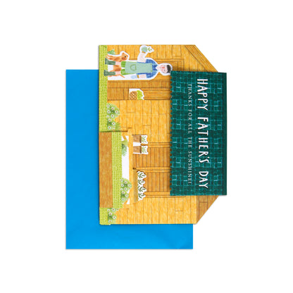 A folded greeting card with a colorful illustrated house scene and a dark green vertical panel reading 'HAPPY FATHER'S DAY' and 'PLANETS OF ALL THE GOURMETS,' shown with a bright blue envelope on a white background.