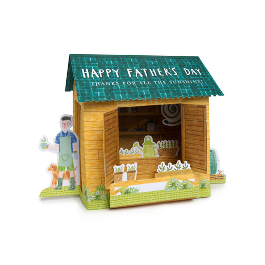 A 3D papercraft model of a wooden garden shed with a green tiled roof and the text 'HAPPY FATHER'S DAY THANKS FOR ALL THE SUNSHINES!' on the roof. The open shed displays a miniature garden interior with plants and a watering can.