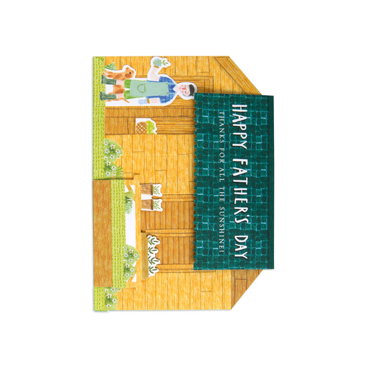 A folded greeting card featuring a hand-drawn wooden cabin illustration with a green vertical banner that reads “HAPPY FATHER’S DAY” and smaller text below, shown on a plain white background.