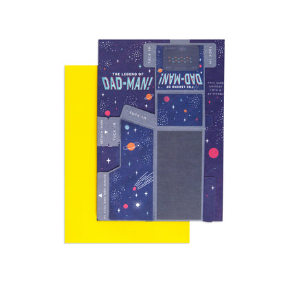 A folded paper card or gift tag with a space-themed design, featuring a dark blue starry background, planets, and the text 'THE LEGEND OF DAD-MAN!' and 'INN-DAD!'. A bright yellow envelope or backing is partially visible behind it on a white background.