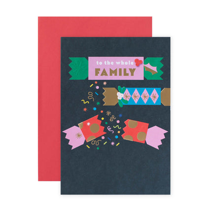 A dark navy greeting card with a colorful Christmas-themed illustration and the words 'to the whole FAMILY' in a pink banner, shown with a red envelope behind it on a white background.