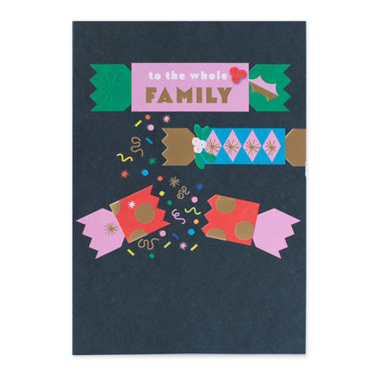 A dark navy blue greeting card featuring whimsical, colorful illustrations of Christmas crackers and festive confetti, with the text 'to the whole FAMILY' on a pink banner near the top.