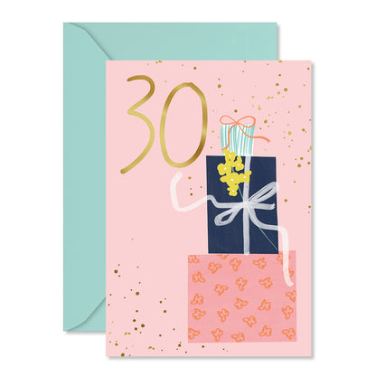 A pink 30th birthday card with gold glitter speckles, a large gold '30' and an illustration of wrapped presents, shown with a light blue envelope behind it on a white background.