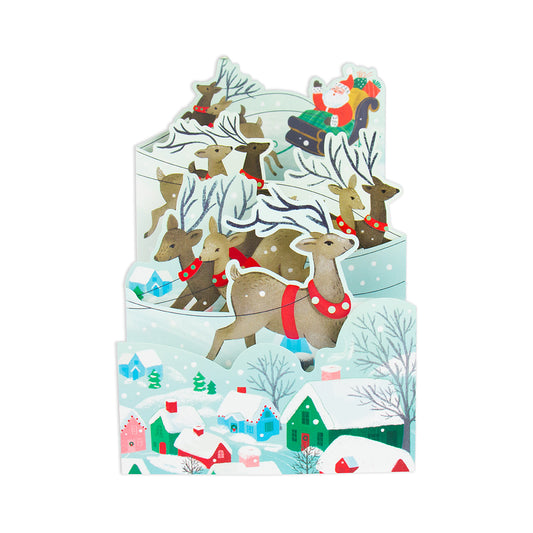 A 3D pop-up cardboard Christmas scene featuring Santa Claus in a sleigh with reindeer, set against a snowy winter village with colorful houses and trees.