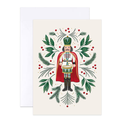 A Christmas greeting card featuring an illustrated nutcracker surrounded by green pine branches, red berries, and delicate gold accents on an off-white background, with a white envelope behind it.
