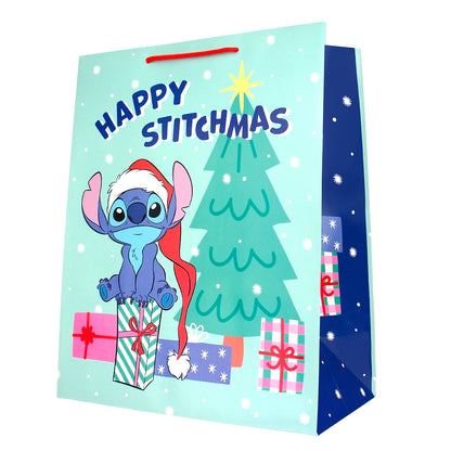 A festive paper gift bag featuring Stitch from Disney's Lilo & Stitch on a light blue holiday background with the text 'Happy Stitchmas,' a green Christmas tree, wrapped presents, and a dark blue side panel with white snowflakes.