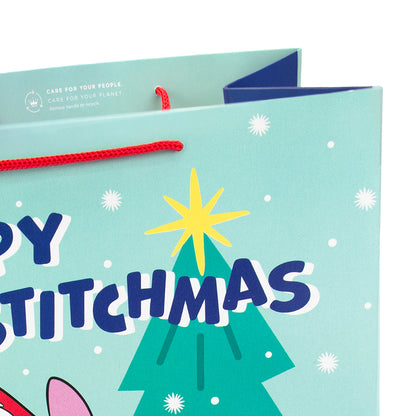 A close-up of a light blue Christmas gift bag with a red rope handle, featuring a cartoon Christmas tree, star, snowflakes, and the text 'HAPPY STITCHMAS' in dark blue and white.
