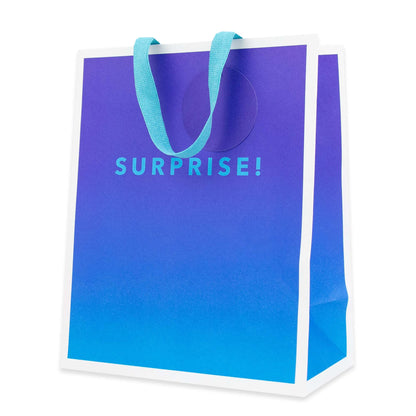 A paper shopping bag with a blue-to-purple gradient front panel, white side borders, and light blue handles. The word 'SURPRISE!' is printed in light blue on the front.