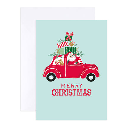 A Christmas greeting card with a light blue background, featuring an illustration of Santa driving a red car loaded with wrapped presents, with the text 'MERRY CHRISTMAS' below. A white envelope is placed behind the card.