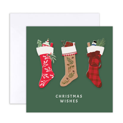 A square Christmas greeting card with a dark green background featuring three illustrated Christmas stockings in red, beige, and plaid, with the words 'CHRISTMAS WISHES' printed below. A white envelope is partially visible behind the card.