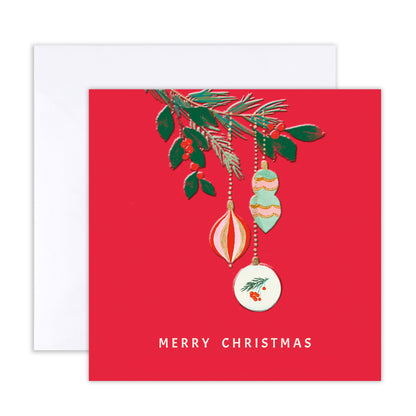 A square Christmas greeting card with a bright red background, featuring an illustrated pine branch with green leaves and hanging ornaments, plus the text 'MERRY CHRISTMAS' at the bottom. A white envelope is partially visible behind the card on a white background.