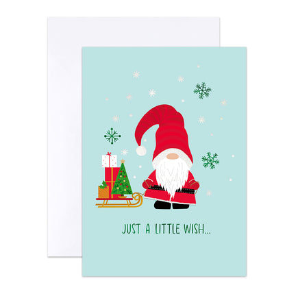 A Christmas greeting card with a light blue background, featuring a cartoon gnome in a red hat and coat beside a sleigh with wrapped gifts and a small tree. The card reads 'JUST A LITTLE WISH.