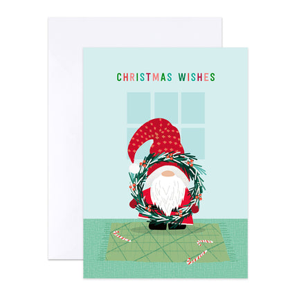 A Christmas greeting card with a white envelope behind it. The card features a cartoon gnome wearing a red hat and holding a green wreath, with the text 'CHRISTMAS WISHES' above.