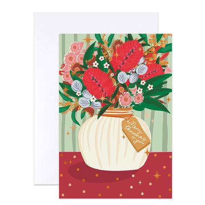 A greeting card with a floral illustration of red broomflowers in a cream vase, with a hanging tag reading 'Merry Christmas you', shown with a white envelope behind it on a white background.