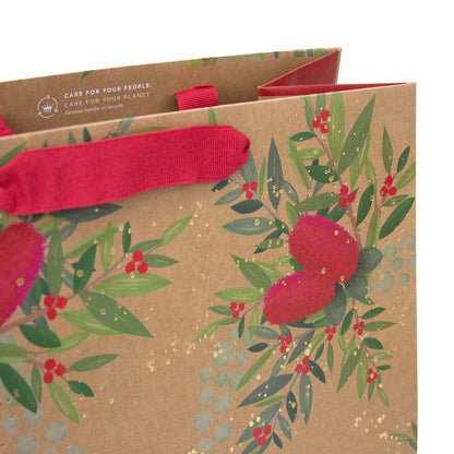A kraft paper gift bag with a red ribbon handle, decorated with green leaves, red berries, and gold glitter, with the text 'CARE FOR YOUR PEOPLE, CARE FOR YOUR PLANET. Non-toxic handle & recycle.