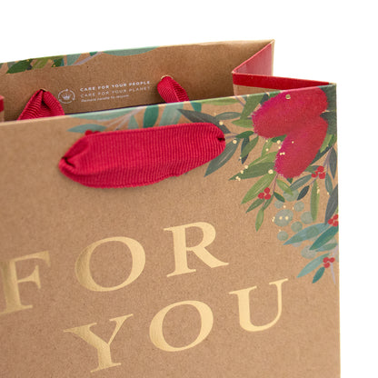A brown kraft paper gift bag with red ribbon handles, decorated with green leaves and red flowers, featuring the text 'FOR YOU' in gold foil on a white background.