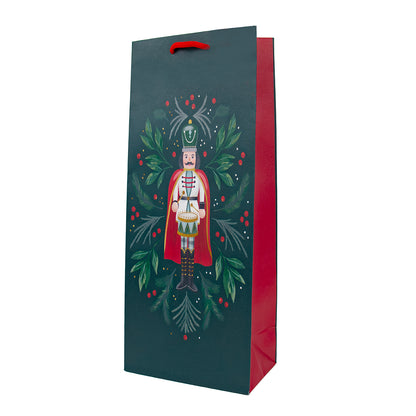 A dark teal Christmas-themed gift bag with a red handle and red inner side, featuring a printed illustration of a nutcracker surrounded by green holly leaves and red berries, shown on a plain white background.