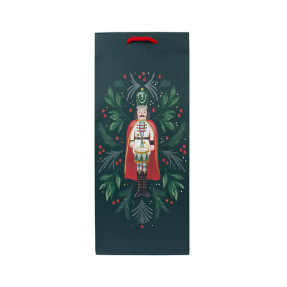 A dark teal Christmas-themed gift bag with a red handle, featuring an illustration of a nutcracker surrounded by green holly leaves and red berries, centered on a plain white background.