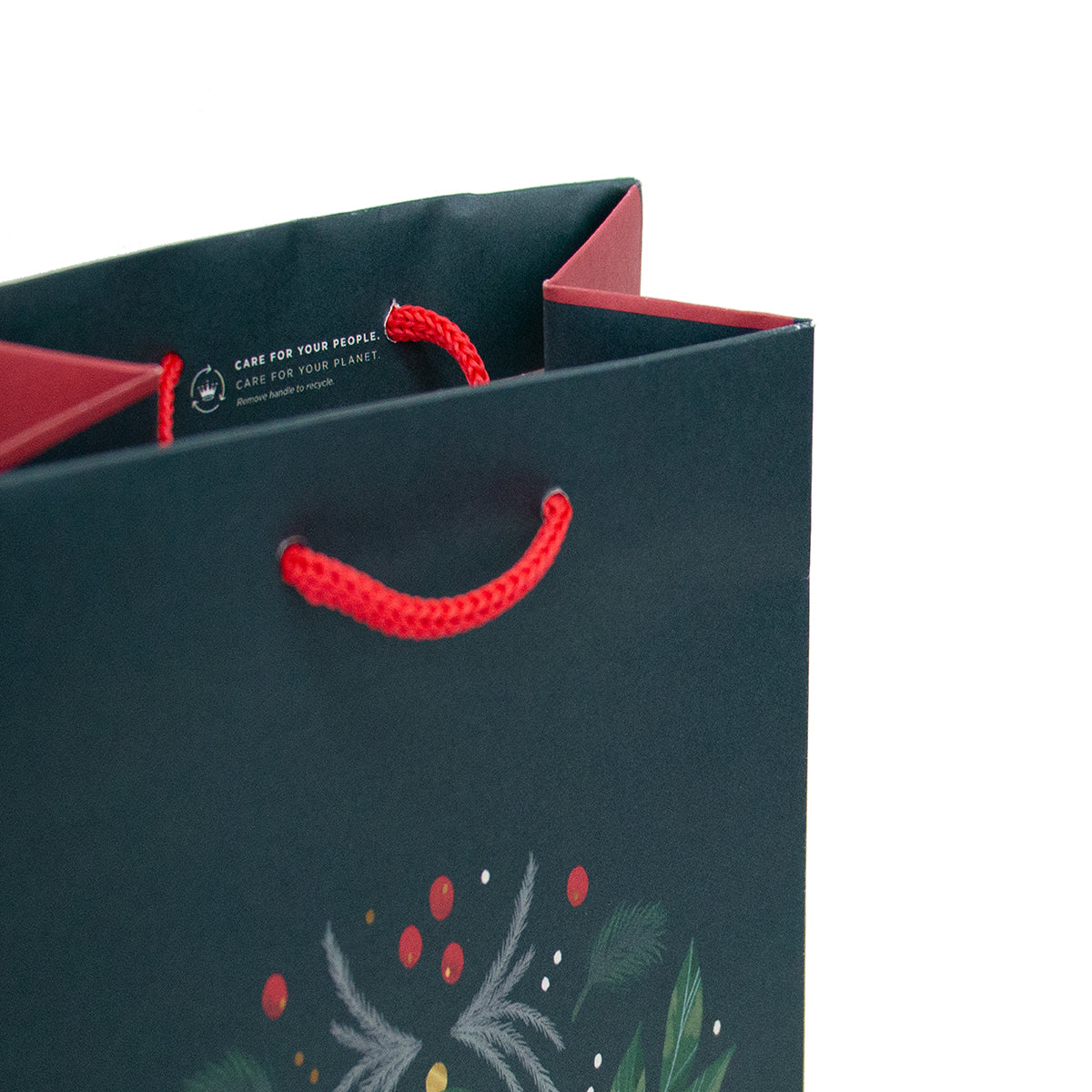 A dark green paper gift bag with red rope handles and a festive holly and pine illustration near the bottom, photographed on a white background.