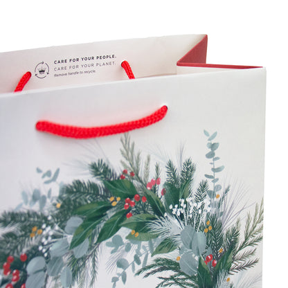 A white paper gift bag with red twisted handles and a printed winter botanical design featuring green pine branches, eucalyptus leaves, and small red berries. The inside top flap shows the text 'CARE FOR YOUR PEOPLE.