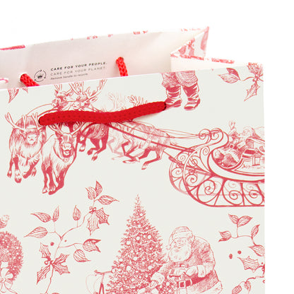 A close-up of a Christmas-themed paper gift bag with red printed illustrations of Santa Claus, reindeer, sleigh, and evergreen tree on an off-white background, with red rope handles and a small printed brand mark near the top.
