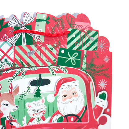 A close-up of a festive Christmas gift card or paper decoration featuring Santa Claus in a red truck, with a deer, a white dog, wrapped presents, pine branches, snowflakes, and red ribbon bows on a white background.