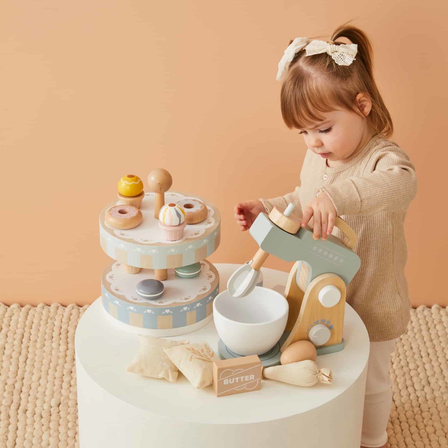 Wooden cake stand set featuring a young girl mixing play food with a realistic mixer on a white table.