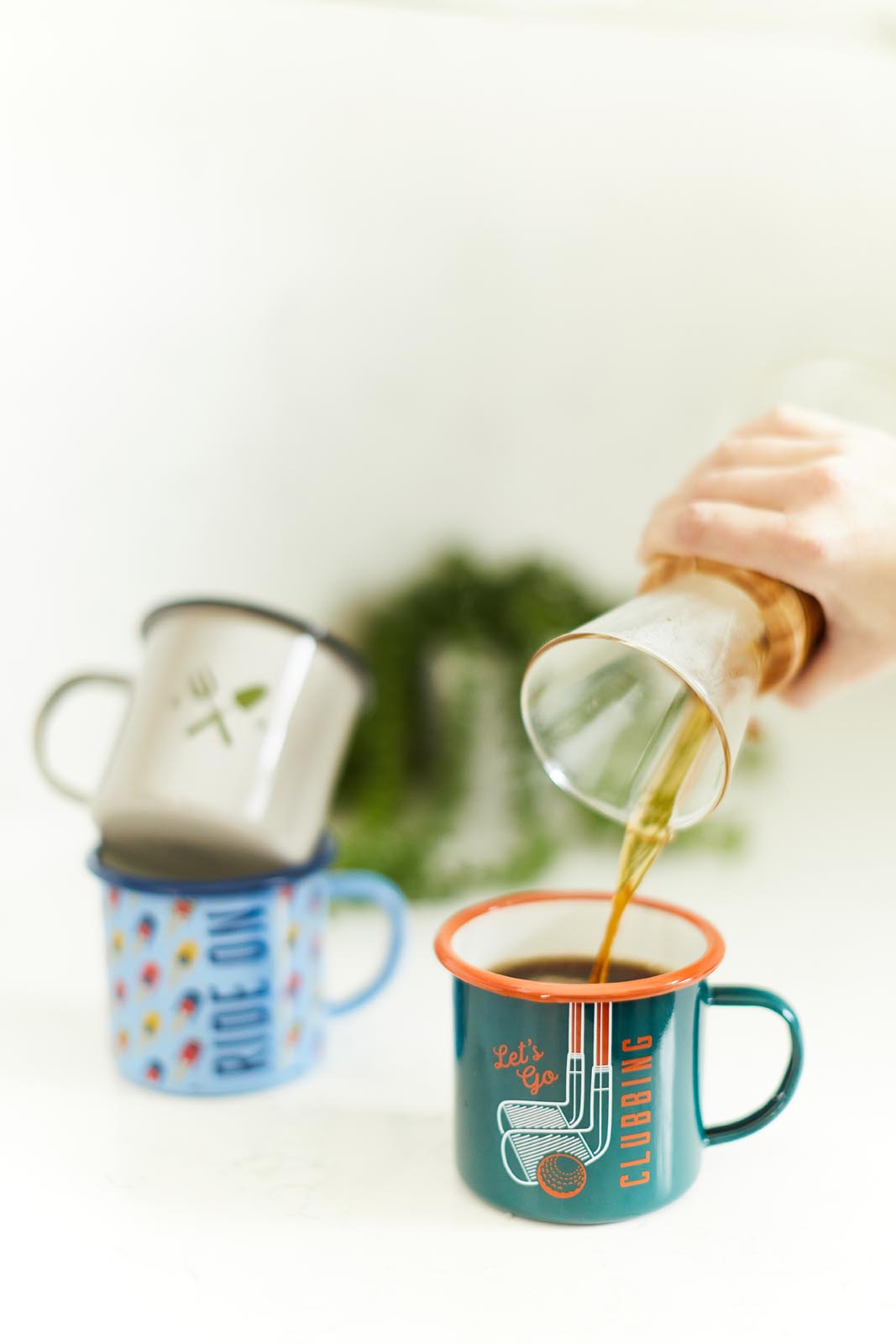 Enamel Mug - Cycling in green with orange rim, being filled with hot drink, with other patterned mugs in background.